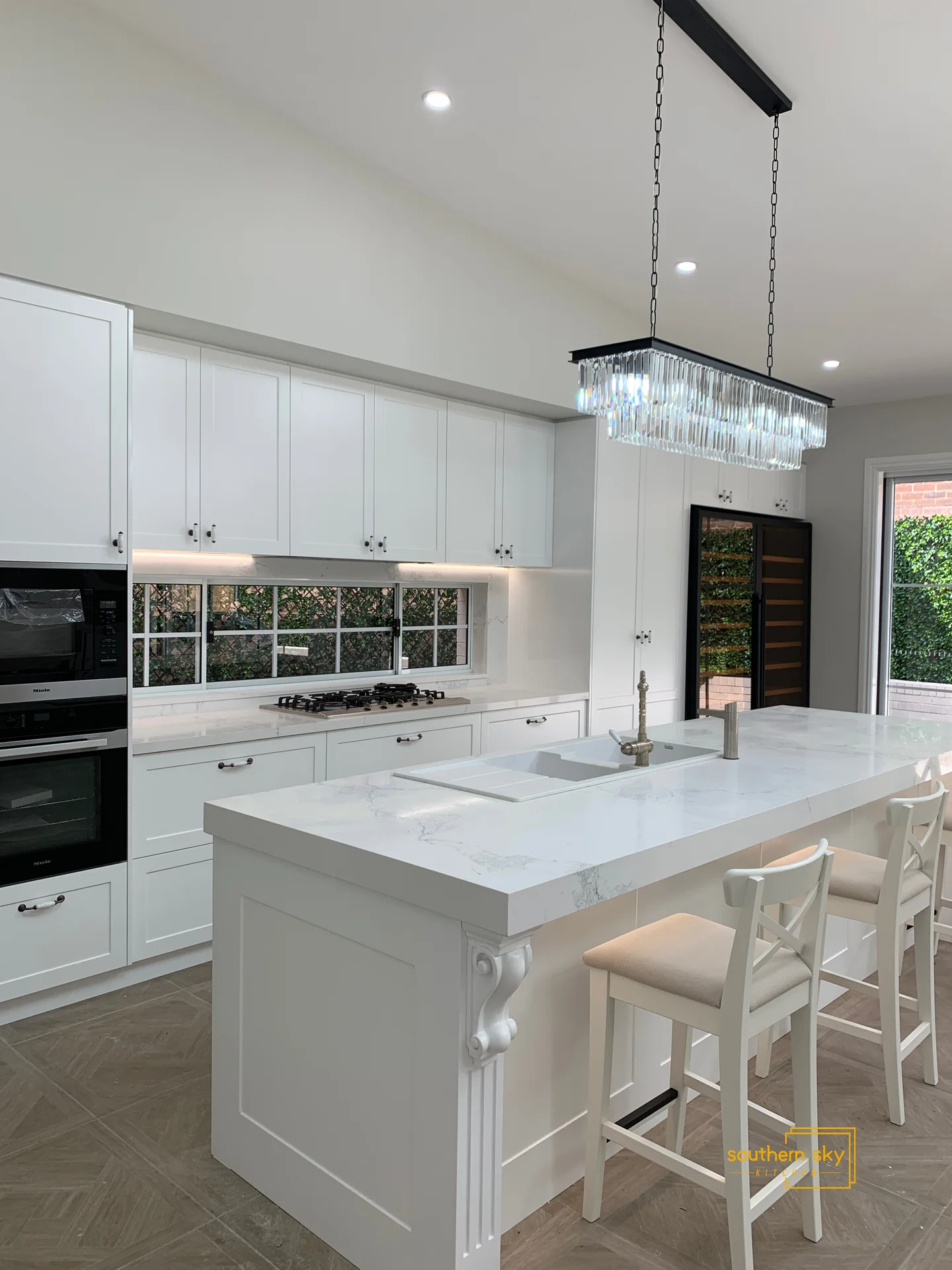 Elegant kitchen with pristine white cabinetry, marble countertops, and a sophisticated chandelier, showcasing exceptional design finesse.